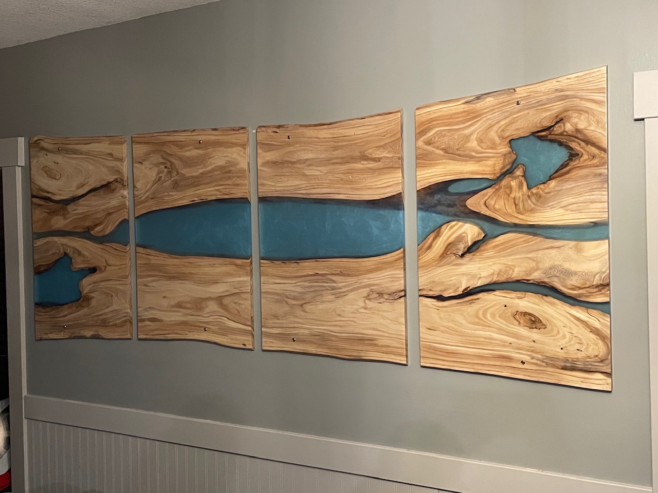 Four-panel live-edge wall installation with a continuous teal epoxy river flowing across the panels, by Lost Species